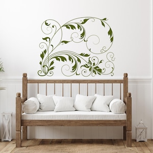 May include: A decorative wall decal featuring a green floral design with leaves and swirls. The decal is placed above a wooden bench with white cushions. The room has a white door and a potted plant.