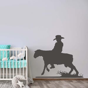 May include: A dark gray wall decal featuring the silhouette of a person wearing a cowboy hat while riding a horse. The design shows the horse in motion with a splash of dirt or dust at its feet, creating a dynamic scene applied to a flat surface.