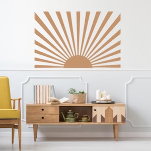 May include: A wall decal featuring a stylized sunburst design in shades of brown. The sun's rays emanate from a semi-circular base, creating a retro aesthetic. The decal is mounted on a white wall above a wooden cabinet.