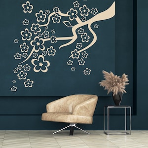 May include: A beige armchair with a silver base sits in front of a dark teal wall. A beige floral branch design decorates the wall. A black vase with dried flowers sits on a small side table.