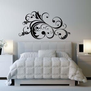 May include: A black floral wall decal above a bed with white bedding. The decal features swirling lines and leaf-like shapes. The room has white bedside tables with lamps on either side of the bed.