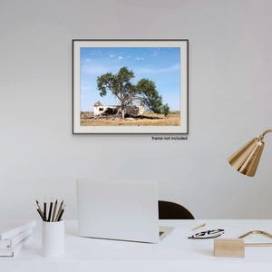 May include: A framed print showing a dilapidated building and a large tree against a blue sky. The artwork hangs on a white wall above a desk with a laptop, lamp, and stationery. The text "frame not included" is visible.