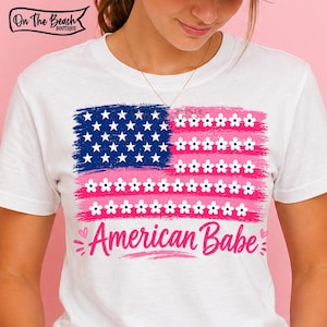 May include: White t-shirt featuring a stylized American flag design. The flag incorporates blue, pink, and white elements, with stars and floral accents. The text "American Babe" is written in pink script below the flag.