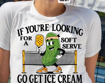 May include: White t-shirt featuring a green pickle character holding a yellow paddle and the text "If you're looking for a soft serve, go get ice cream".