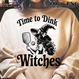 May include: Cream-colored sweatshirt featuring a black and white graphic of a witch playing pickleball. The text "Time to Dink Witches" is above and below the illustration. The design includes a witch's hat, paddle, and ball.