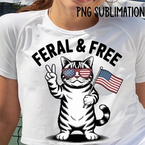 May include: White t-shirt featuring a black and white illustration of a cat wearing sunglasses with an American flag design and holding a small American flag. The text "FERAL & FREE" is above the cat.