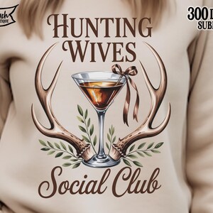 May include: A beige sweatshirt with a graphic featuring a martini glass, antlers, and the text "Hunting Wives Social Club." The design includes brown and green accents, and the words "300 DPI PNG Sublimation" are also visible.