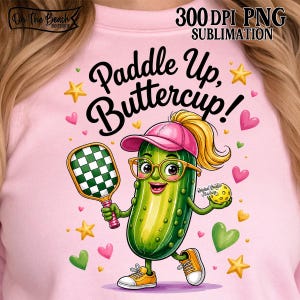 Op de afbeelding: Een lichtroze sweatshirt met een cartoon augurkfiguur met een roze pet en een bril. De augurk houdt een groen-wit geblokt pickleball-racket en een gele bal vast. De tekst luidt Paddle Up, Buttercup! met sterren en hartjes in de afbeelding.