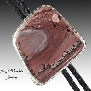 May include: A bolo tie featuring a large, rectangular stone with a silver-colored border. The stone has a marbled appearance with shades of red, pink, and white. The black leather cord is braided. The text "Stony Mountain Jewelry" is in the lower left corner.