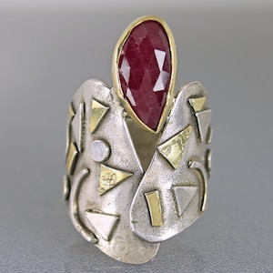 May include: A wide silver ring with gold accents and a large, teardrop-shaped, faceted ruby gemstone. The ring features geometric shapes and a textured surface, creating a bold and artistic statement piece.