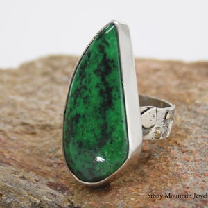 May include: A large, silver ring featuring a vibrant green stone with black speckles. The stone is set in a polished silver bezel, with a textured band and small gold accents. The ring is a statement piece of jewelry.