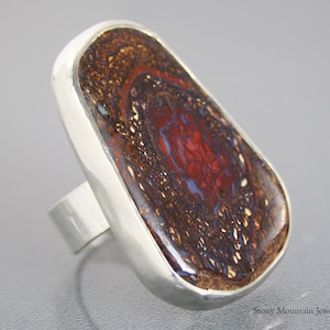 May include: A large, oval-shaped ring featuring a polished, multi-colored stone with red, brown, and blue hues. The stone is set in a wide, silver-colored band. The ring has a modern, bold design, suitable for statement jewelry.