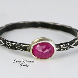 May include: A silver ring with a textured band and a faceted, oval-shaped ruby gemstone. The ring is a delicate piece of jewelry, perfect for everyday wear. The text "Stony Mountain Jewelry" is visible.