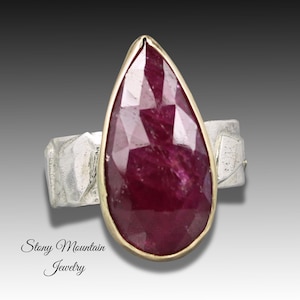 May include: A teardrop-shaped ruby gemstone ring with a gold-colored bezel and a silver-colored band. The ruby is a deep red color and has a faceted cut. The ring is set against a gradient background.