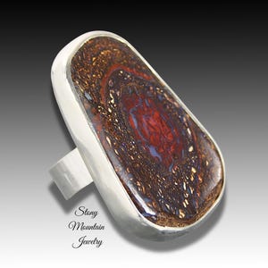 May include: A silver ring featuring a large, irregular, polished boulder opal gemstone with deep red, brown, and metallic gold inclusions. The stone is set in a smooth silver bezel. The text Stony Mountain Jewelry is printed in black.