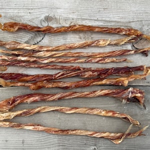 May include: A collection of twisted, dried dog treats. The treats are a mix of light tan and reddish-brown, with a textured, braided appearance. They are arranged on a weathered, gray wooden surface.