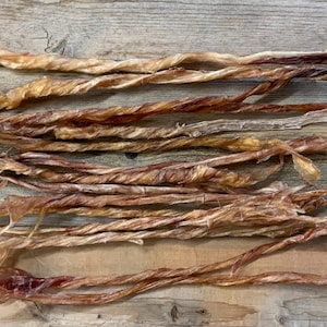 May include: A collection of dried, twisted dog treats, in shades of brown and tan, are arranged on a wooden surface. The treats have a textured appearance and vary in thickness and length, suggesting a natural product.