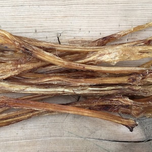 May include: A pile of dried, light brown dog treats, resembling thin, elongated strips. The treats have a slightly translucent appearance and are arranged on a light-colored wooden surface. The texture appears to be rough and natural.