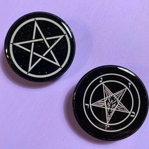 May include: Two black, round pins with white pentagram designs. One pin features a simple five-pointed star, while the other displays a star with a goat-like face inside. Both have a glittery texture.