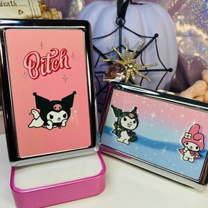 Op de afbeelding: Twee roze metalen koffers met cartoonkarakterontwerpen. E&eacute;n koffer heeft het woord "Bitch" in rode letters en een zwart cartoonkarakter. De andere koffer heeft een ander cartoonkarakterontwerp. Een witte pompoen en een spinnenwebdecoratie staan op de achtergrond.