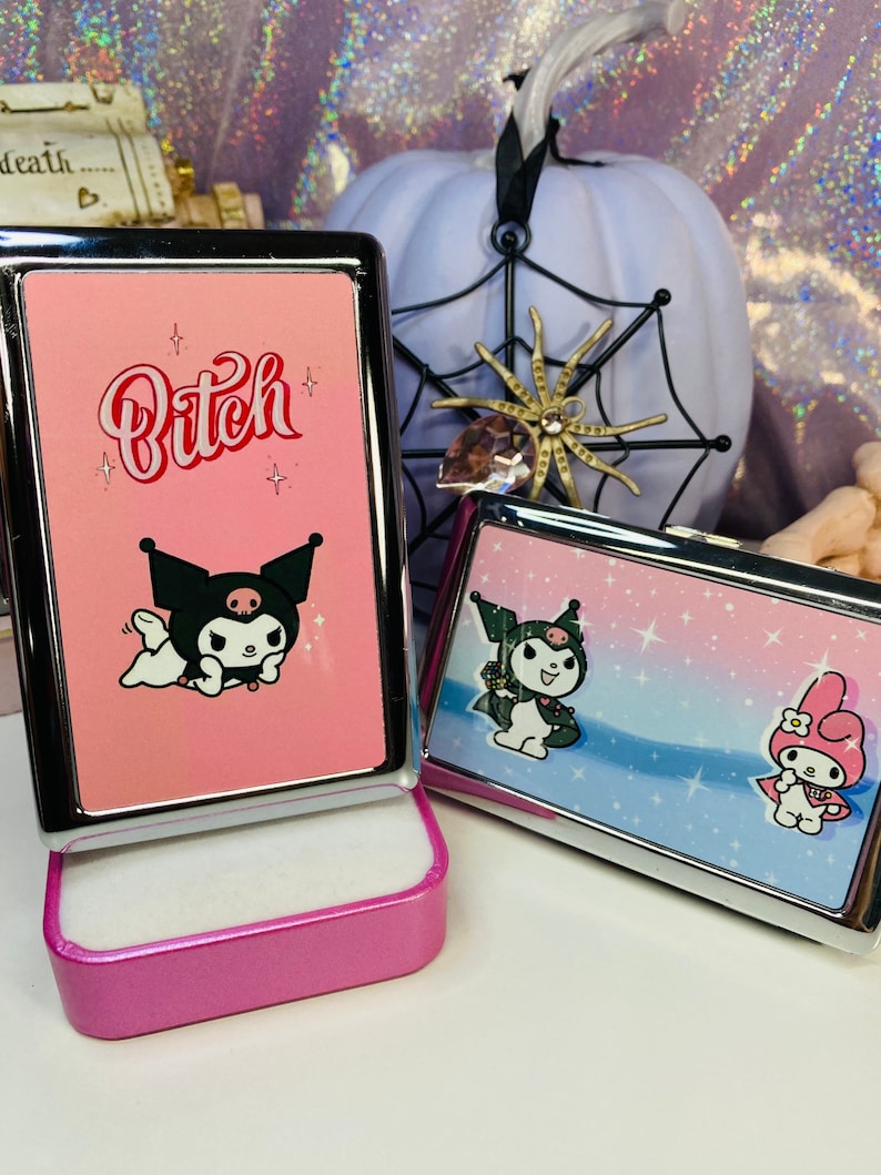 Op de afbeelding: Twee roze metalen koffers met cartoonkarakterontwerpen. E&eacute;n koffer heeft het woord "Bitch" in rode letters en een zwart cartoonkarakter. De andere koffer heeft een ander cartoonkarakterontwerp. Een witte pompoen en een spinnenwebdecoratie staan op de achtergrond.