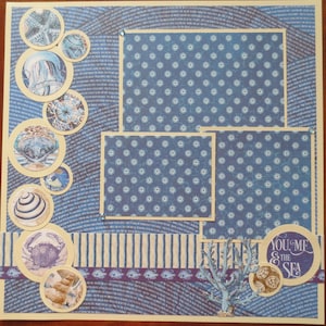May include: A scrapbook page with a nautical theme. It features blue patterned paper, circular cutouts with sea creature illustrations, and rectangular photo mats. The phrase "YOU ME & THE SEA" is displayed on a circular label, with a coral and seashell embellishment.