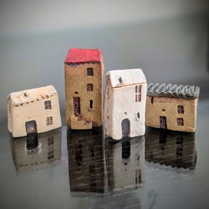 May include: Four miniature ceramic house figurines with textured surfaces. One tall house has a red roof, while the others are beige or white with small windows and doors. The structures feature varied architectural details and earthy tones.