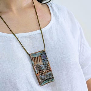 May include: A rectangular ceramic pendant featuring a textured, multi-colored glaze in shades of blue, copper, and brown with linear embossed patterns. The pendant is suspended from a thin, dark olive green cord that rests against a white fabric.