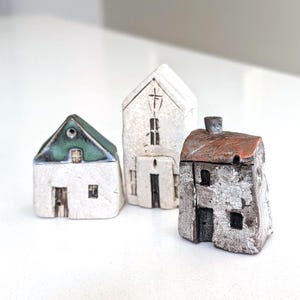 May include: Three small ceramic house figurines. One has a white base and green roof, one is a tall white structure with a cross, and one has a textured grey base with a brown roof. Each piece features hand-painted windows and doors.