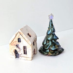 May include: A small ceramic house with a textured off-white finish, dark windows, and a door, positioned next to a conical ceramic Christmas tree with a glossy, iridescent dark green glaze and a clear, star-shaped topper with a purple tint.