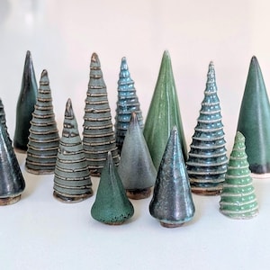 May include: A collection of fifteen ceramic tree figurines in various conical shapes and sizes. The trees feature diverse finishes, including smooth solid green glazes, textured horizontal ridges, and mottled blue-grey tones with glossy surfaces.