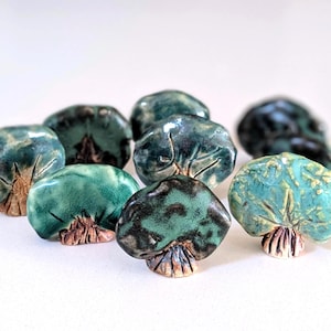 May include: A collection of small, mushroom-shaped ceramic knobs featuring a glossy, mottled glaze in shades of teal, turquoise, and dark green, with a textured, light brown base that resembles a stalk, arranged in a cluster on a white surface.