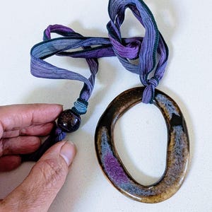 Può includere: Collana artigianale con un grande pendente ovale con finitura lucida nei toni del marrone, viola e blu. Il pendente è sospeso a un nastro viola e blu. Una perla di legno scuro adorna il nastro.