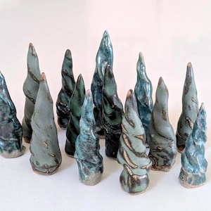May include: A collection of thirteen ceramic sculptures shaped like twisted, conical trees. Each piece features a glossy glaze in varying shades of teal, sage green, and dark charcoal, with textured, spiraling surfaces and pointed tops.
