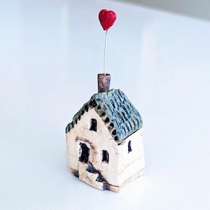 May include: A small ceramic house figurine with a cream-colored base and a textured blue-grey roof. A thin metal wire extends from the chimney, topped with a small, bright red heart. The structure features carved windows and a dark doorway.