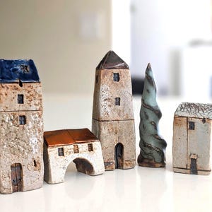 May include: A collection of five ceramic miniature structures including three tall buildings with textured beige walls, one low arched building with a copper-colored roof, and one tall, spiraling, sage green tree-shaped sculpture.