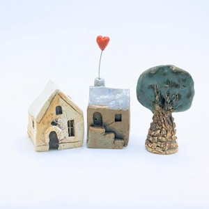 Puede incluir: Un conjunto de tres esculturas de cerámica en miniatura que consta de dos casas pequeñas y un árbol. Una casa es de color crema con techo a dos aguas, la otra es de color canela con techo plano y un corazón rojo en un alambre, el árbol tiene un tronco marrón.