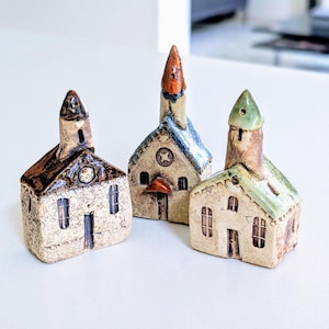 Pode incluir: Três pequenas figuras de casas em cerâmica com torres pontiagudas. A casa da esquerda tem um telhado castanho escuro, a do centro um telhado azul com um detalhe em cobre e a da direita um telhado verde claro. Cada peça tem janelas.