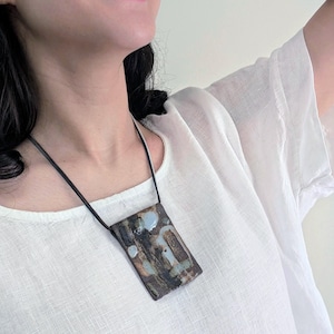 May include: A rectangular ceramic pendant necklace with a black leather cord. The pendant features a mottled design with shades of brown, blue, and white. The necklace is worn against a white shirt.