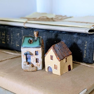 May include: Two ceramic miniature houses. One is off-white with a green roof and blue door. The other is tan with a brown roof and red door. Both have small windows. They are placed on a brown surface with books in the background.