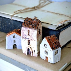 May include: Three miniature ceramic houses with textured white walls and glossy brown roofs. The tallest house features a chimney and multiple windows, while the two smaller houses have arched doorways and detailed window cutouts in their facades.