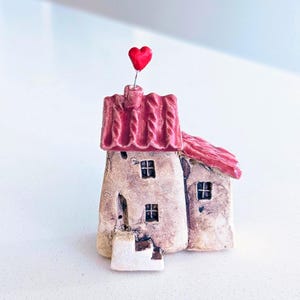 May include: A handcrafted ceramic house figurine with a red roof and a small red heart on top. The house has a rustic, textured appearance with small square windows and a door. The figurine is a charming decorative item.