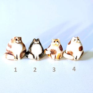 May include: Four small ceramic cat figurines arranged in a row, labeled 1 through 4. Each cat has a rounded body, a white chest, and distinct brown, black, or orange patches. The figures feature simple painted faces and tails.