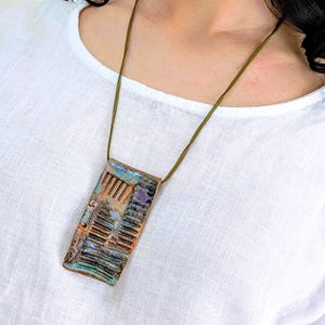 Puede incluir: Un colgante rectangular de cerámica con una superficie texturizada con ranuras verticales y un esmalte multicolor en tonos azul, cobre y morado, suspendido de un cordón fino de color verde oliva sobre una tela de lino blanca.