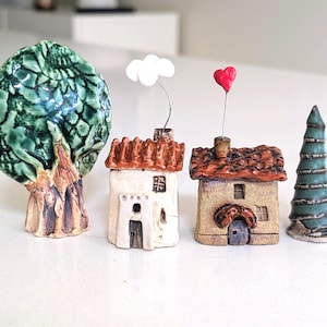 May include: A set of four ceramic figurines including a large green tree with a textured trunk, a white house with a red roof and a white cloud topper, a tan house with a red roof and a red heart topper, and a tall, conical green pine tree.