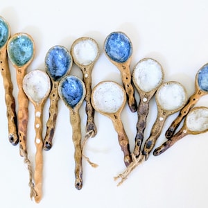 May include: A collection of handmade ceramic spoons with textured, earthy-toned handles and glazed bowls. The bowls feature shades of blue and white. Some spoons have small white dots and twine accents.