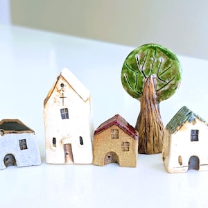 Op de afbeelding: Een verzameling miniatuur keramische huisjes en een boom. De huisjes zijn in verschillende kleuren, waaronder wit, beige en blauw, met details zoals ramen, deuren en een kruis. De boom heeft een groene, bladerrijke top en een bruine stam.