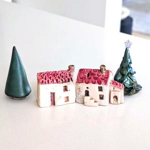 May include: A set of ceramic miniature items including a smooth dark green cone-shaped tree, two small houses with cream-colored walls and textured red roofs, and a dark green textured tree topped with a small purple star-shaped ornament.