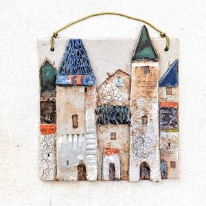 May include: A square ceramic wall hanging featuring a relief of a small village with multiple buildings. The structures have textured white walls, blue and green pointed roofs, and small square windows. A thin brass wire is attached for hanging.