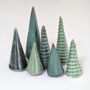 May include: A collection of seven ceramic tree figurines in various shades of green. The set includes smooth conical shapes in dark and light green, alongside textured pieces with horizontal ridges and layered, tiered designs.
